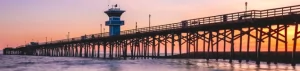 Seal Beach Pier extending over the Pacific Ocean at sunset, with colorful pink and orange skies reflecting across the waves and shoreline.